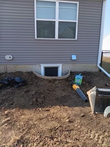 Exterior of a house with a window well, dirt, and gardening tools. Grey siding, light-colored stone base.
