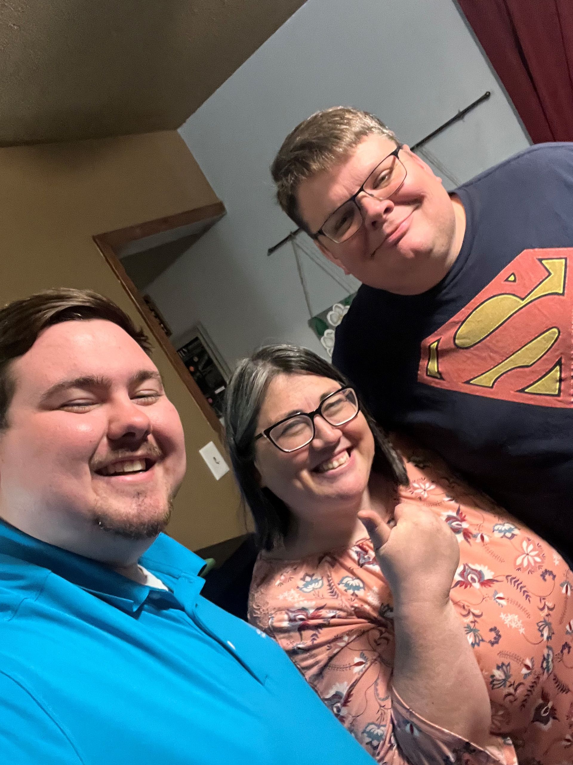 Three smiling people, indoors. Man in blue shirt, woman in floral top, and man in Superman shirt.