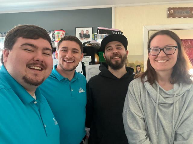 Four smiling people pose for a photo indoors. Two men wear blue shirts, one wears a black hoodie and a cap, and a woman wears a gray sweatshirt and glasses.