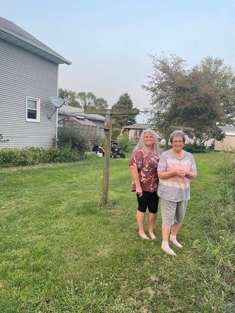 Two barefoot women stand in a grassy yard, smiling. One wears a floral shirt, the other tie-dye. A gray house is in the background.