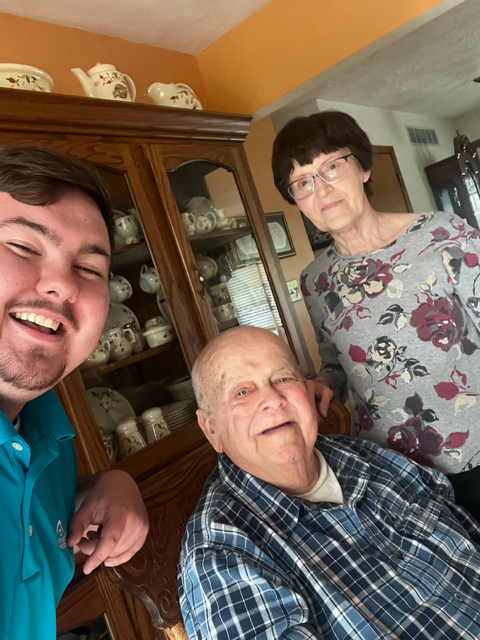 Three people smiling for a selfie; a young man, an elderly man, and an elderly woman, in front of a wooden cabinet.