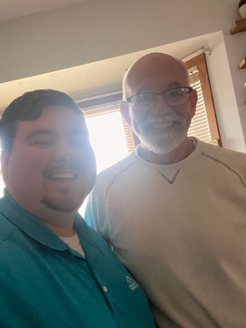 Two men smiling at the camera. One in a turquoise shirt, the other in a white shirt, indoors.