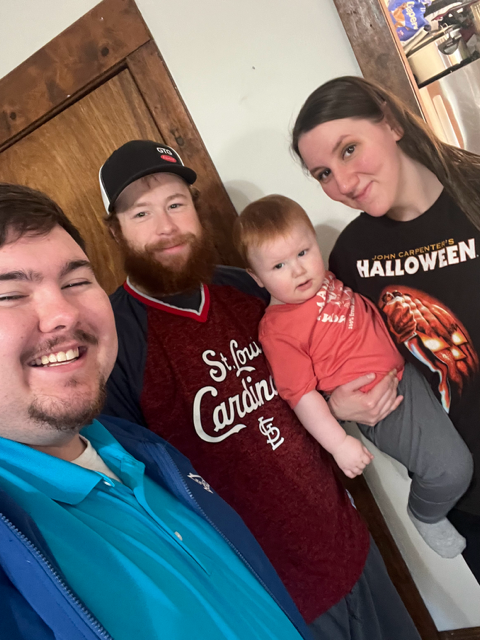 Group of four smiling people: man, man with beard, baby, woman wearing 