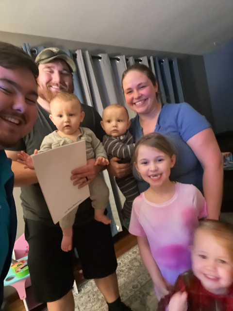 Family smiles, including two babies held by a man, a young girl, and another child, indoors.