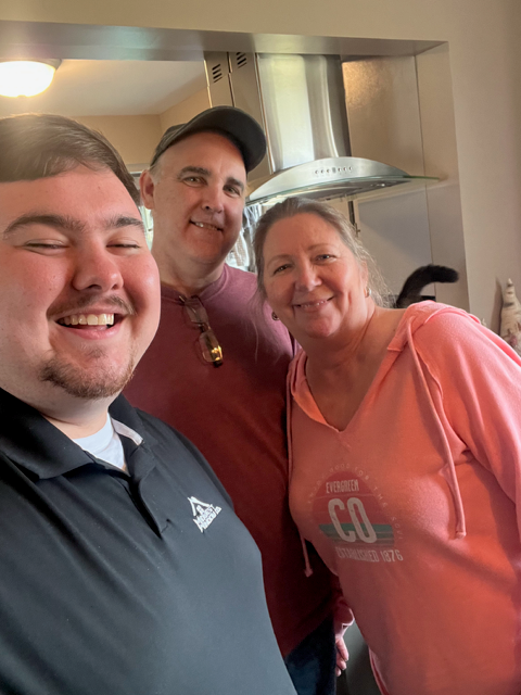 Three people smiling in a kitchen: a young man, a man in a cap, and a woman in pink.