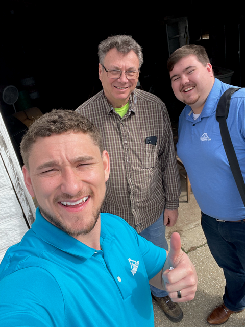Three men smiling, one giving a thumbs-up. Outdoor setting. One in blue, two in casual button-downs.