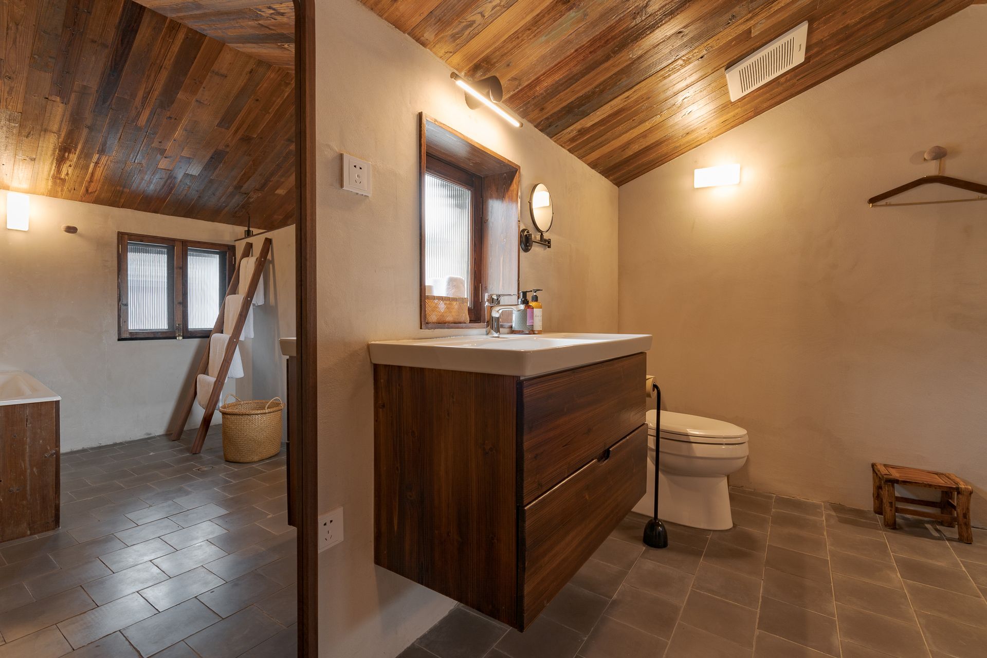 Small rustic bathroom with wood ceiling, beige tile, vanity, mirror, toilet, and window.
