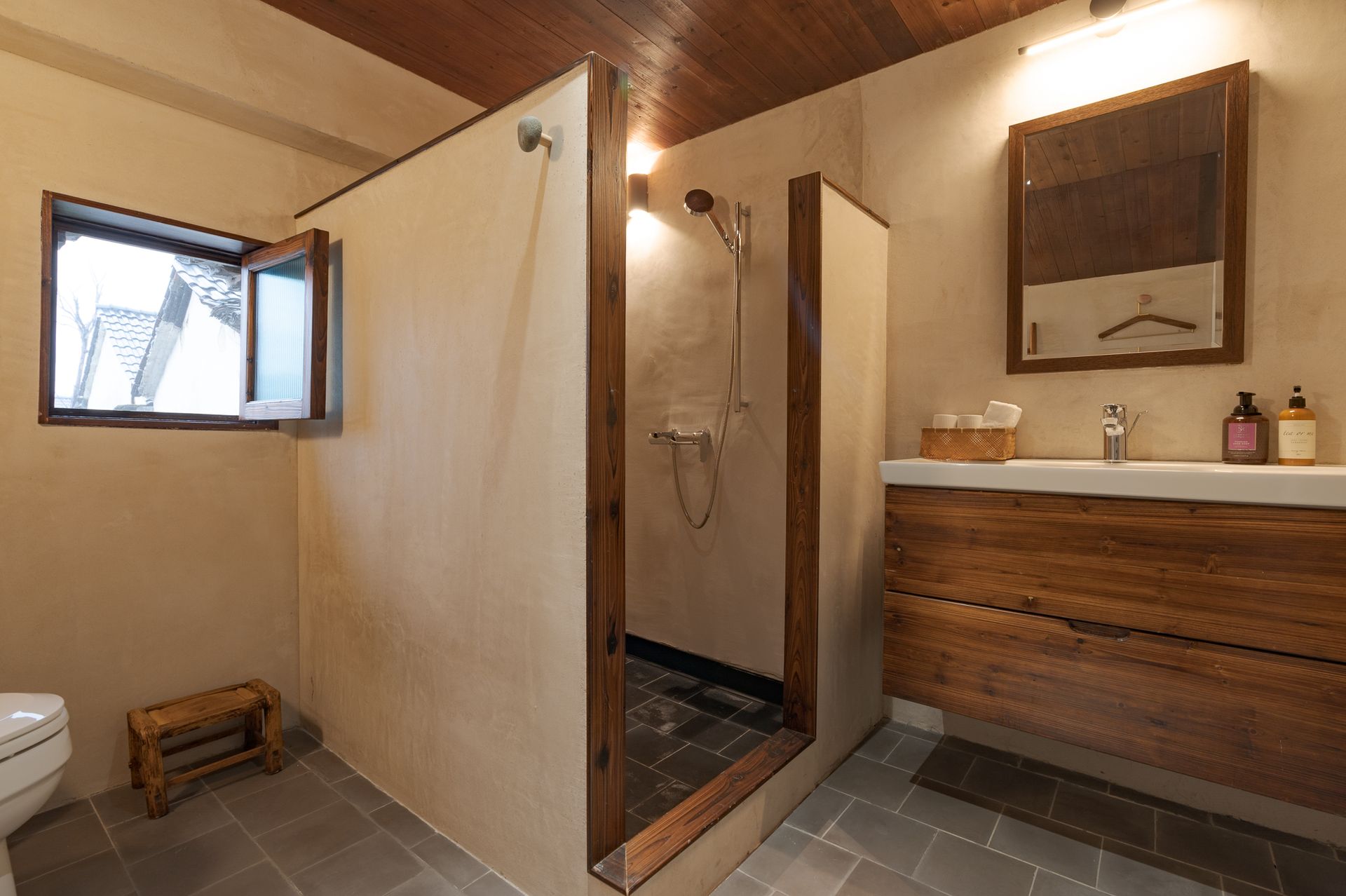 Rustic bathroom with wood vanity, tiled shower, and small window, viewed from the toilet area