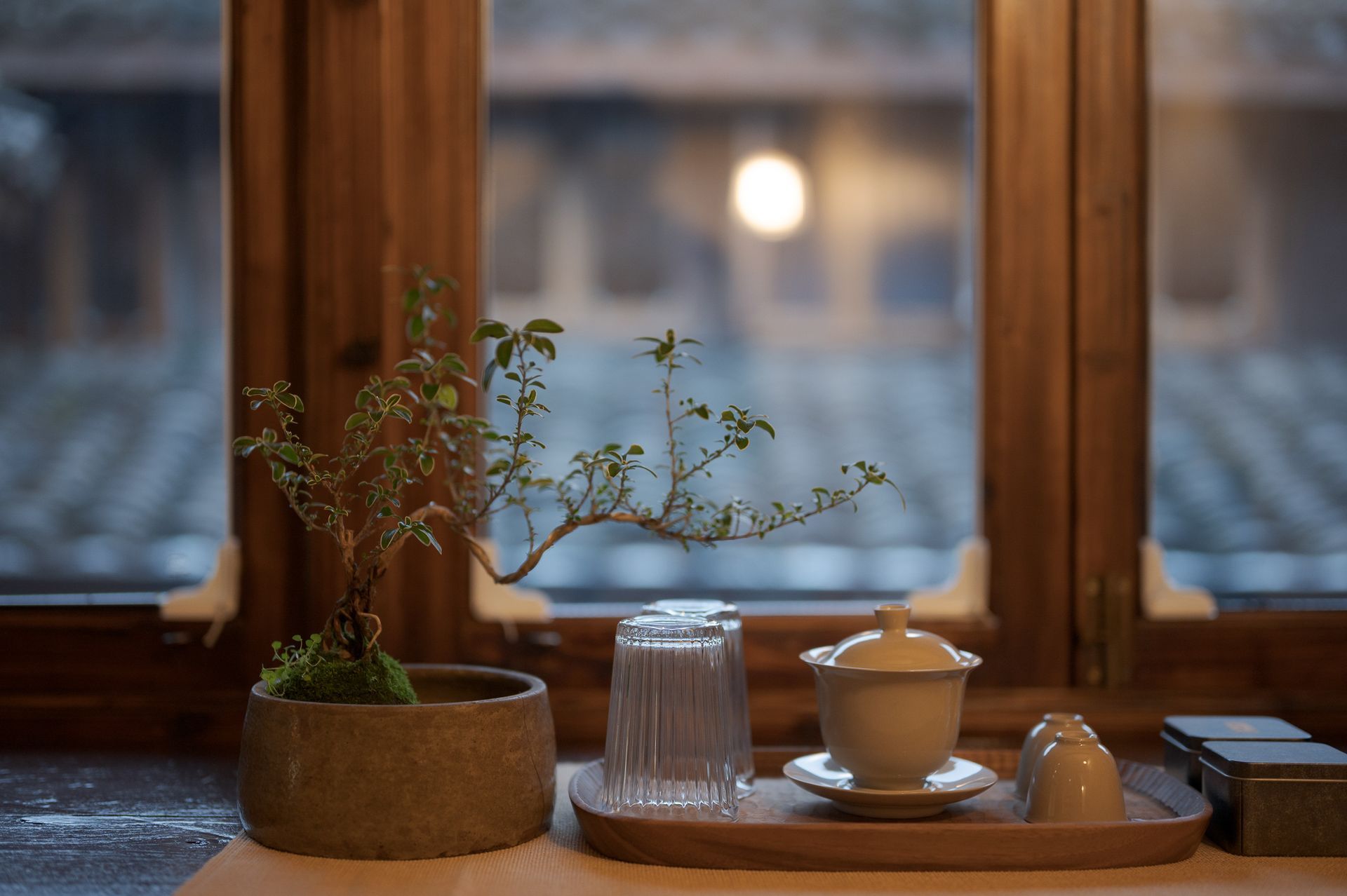 Tea set on a wooden tray by a window, with a potted plant and blurred rooftops outside.