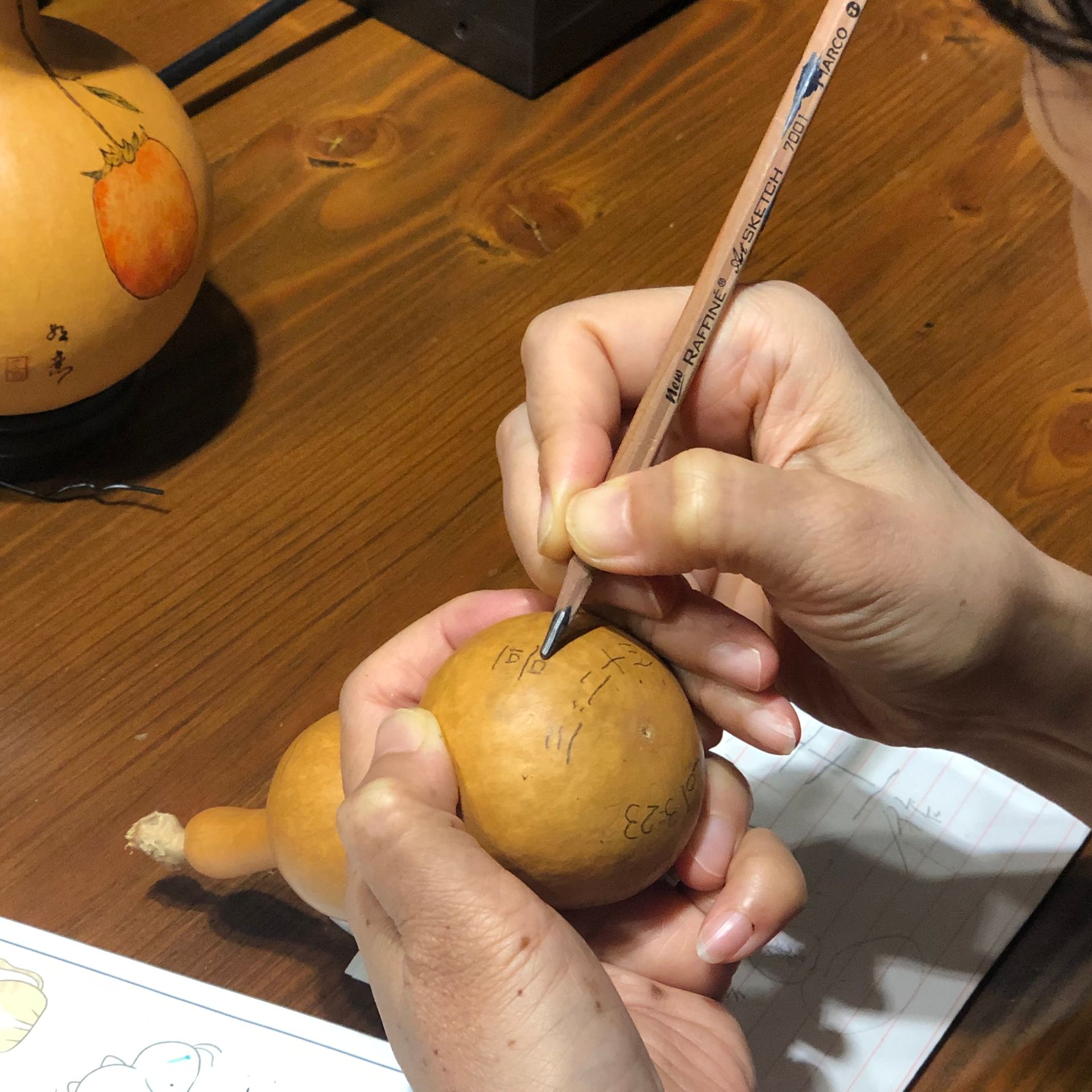 Hands using a pencil to carve or mark a tan fruit on a wooden table