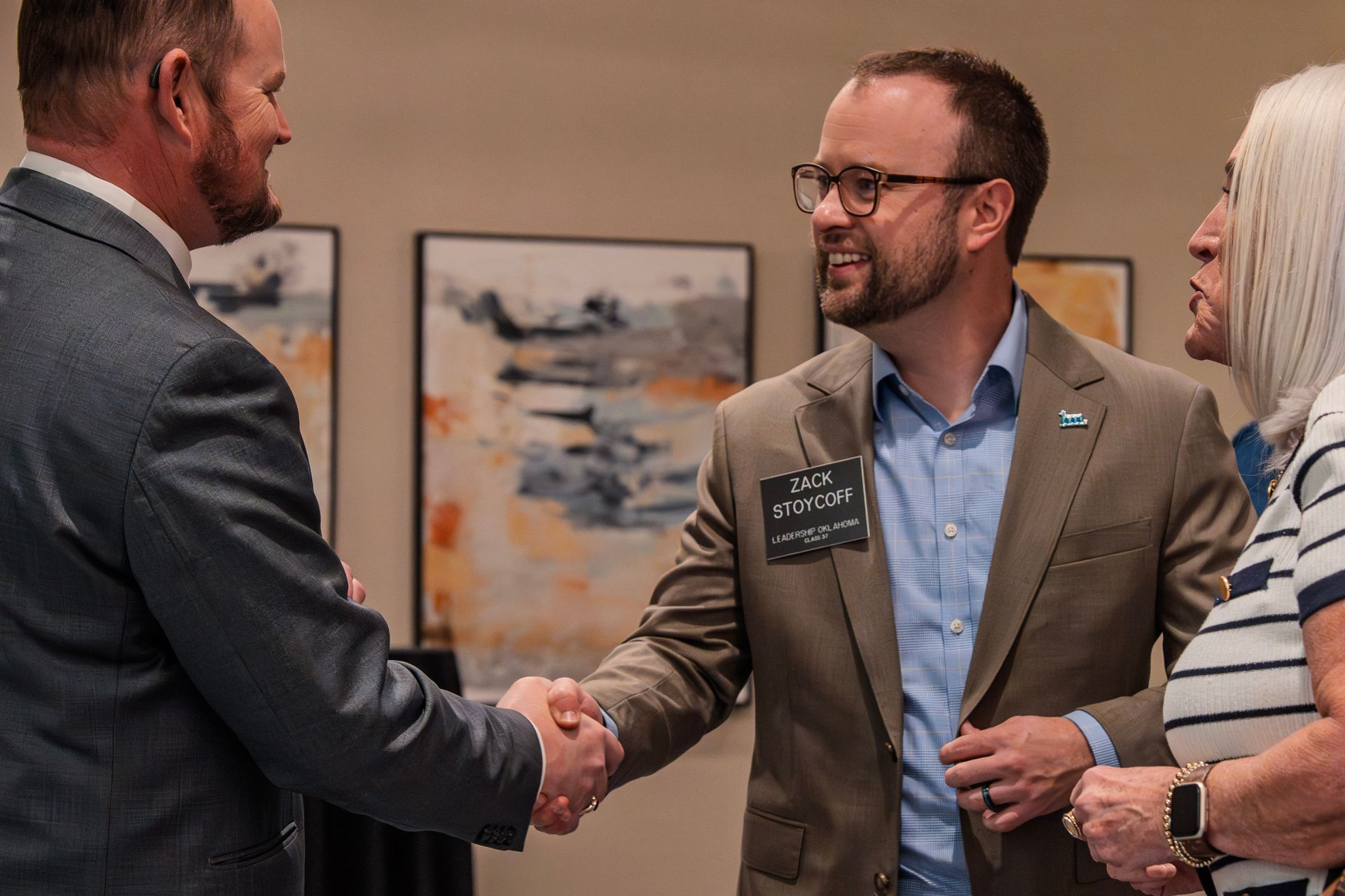 Man in blazer shakes hands with another man, woman watches. Artwork on wall behind.