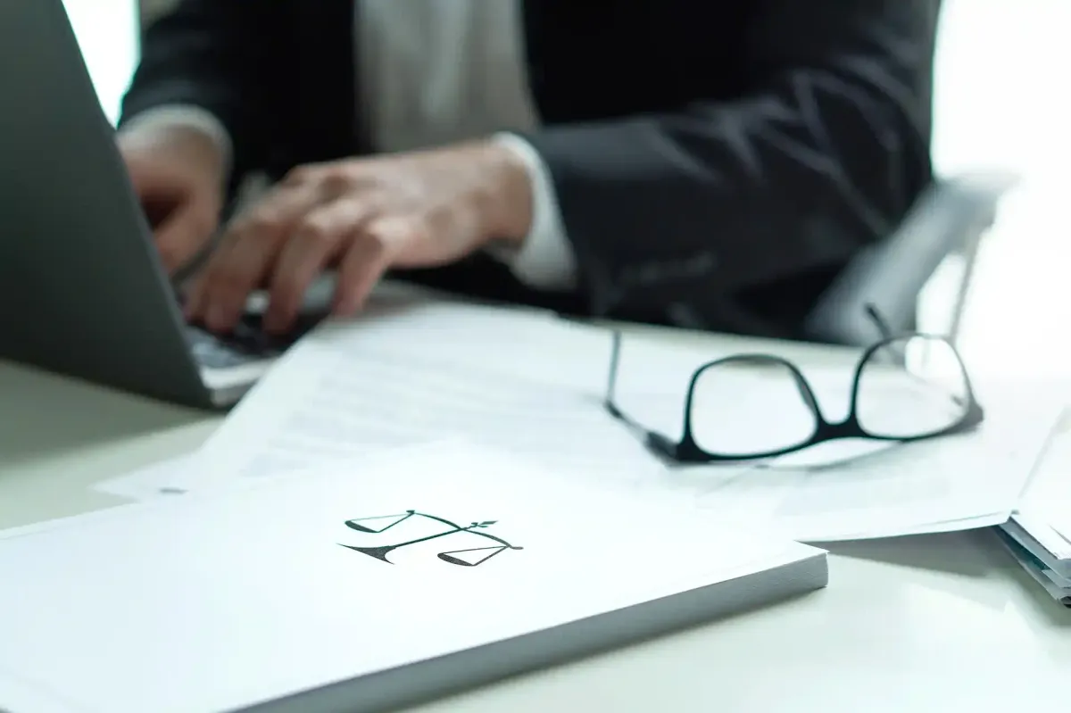 Persona in giacca e cravatta che scrive sul computer portatile, con documenti e occhiali sulla scrivania e un libro di giurisprudenza in primo piano.
