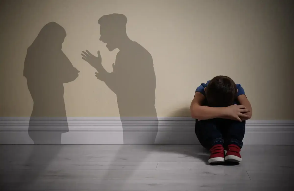 Il bambino si copre il viso sul pavimento; ombre di adulti che litigano sul muro.