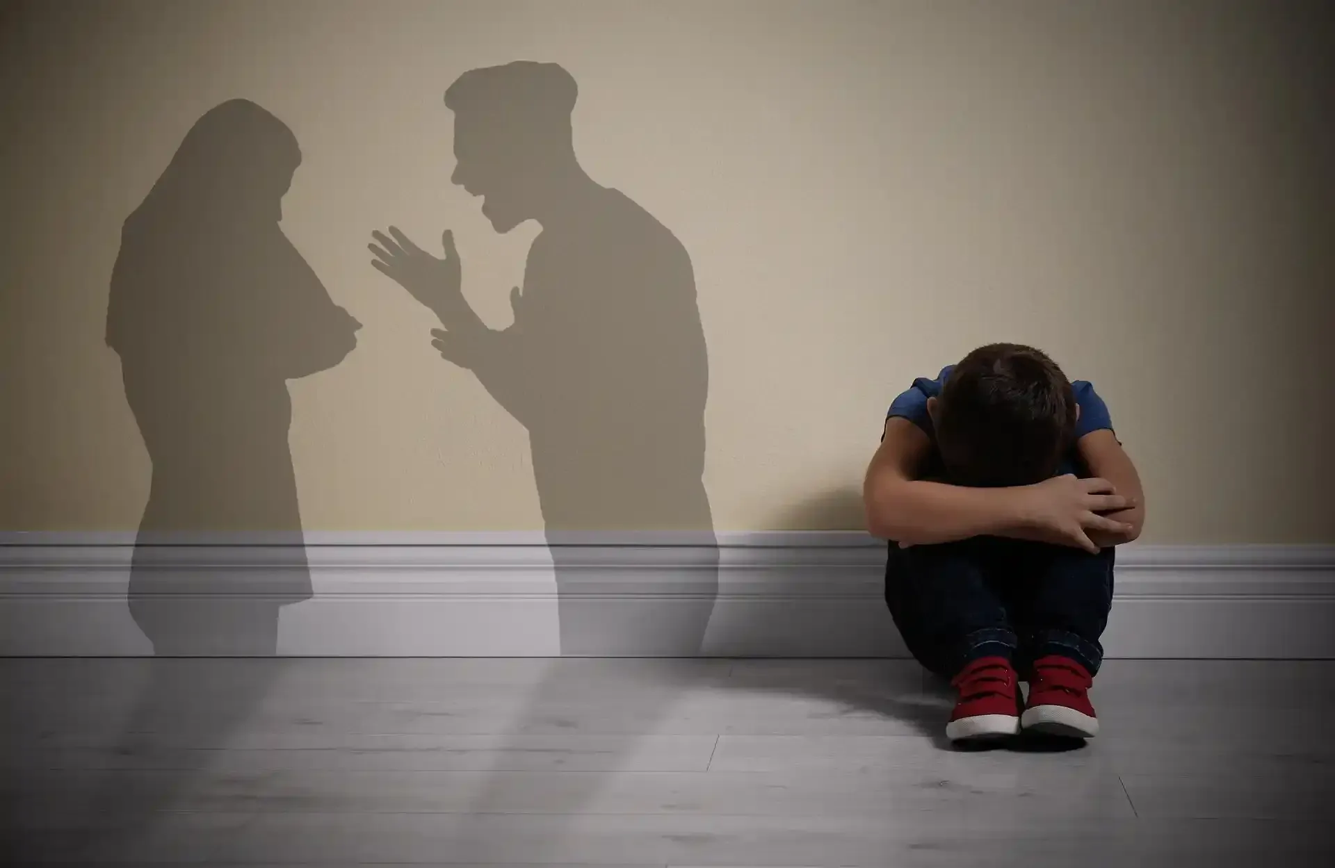 Il bambino si copre il viso sul pavimento; ombre di adulti che litigano sul muro.