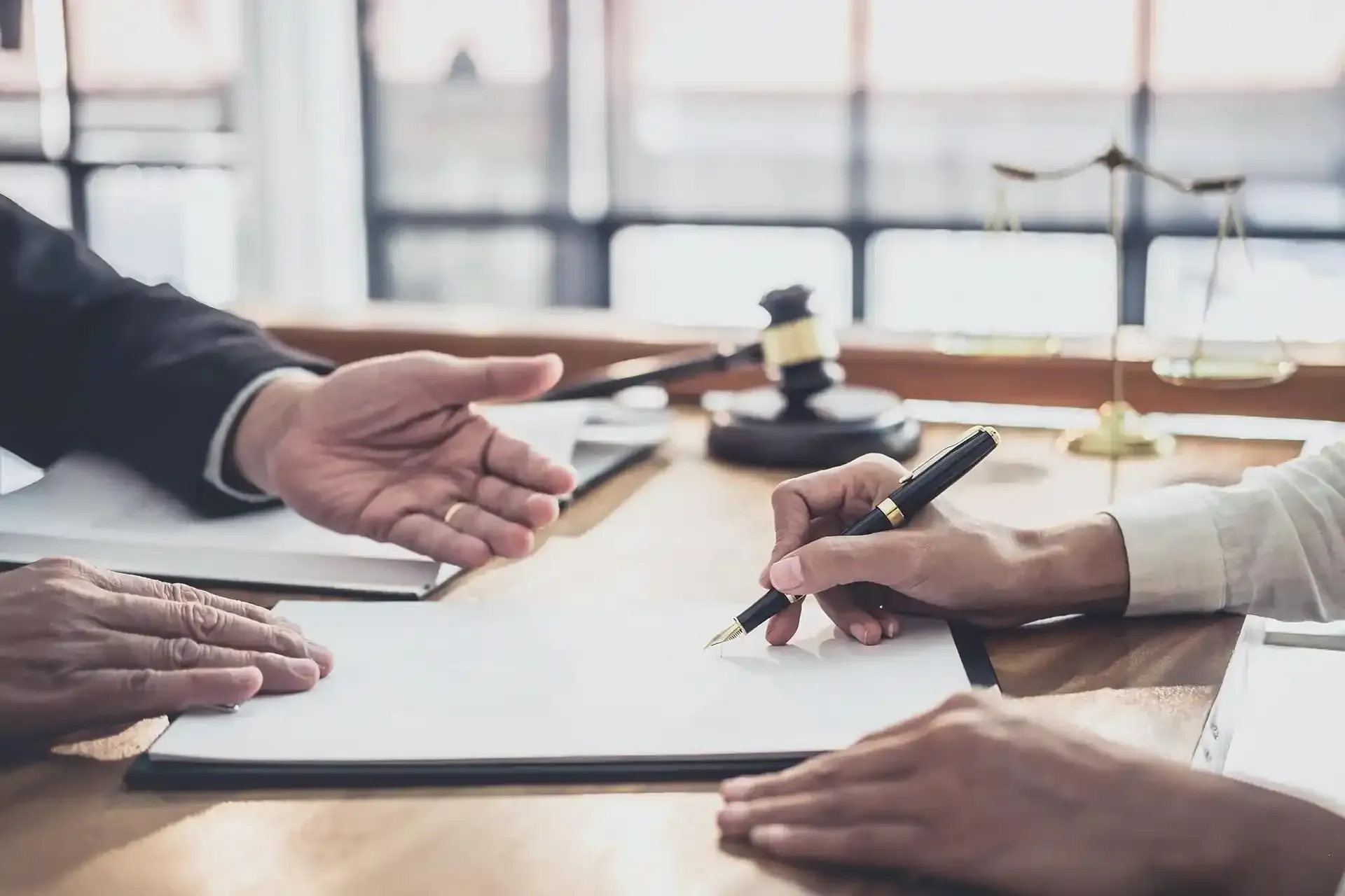 Mani che firmano un documento su una scrivania, martelletto e bilancia della giustizia sullo sfondo.
