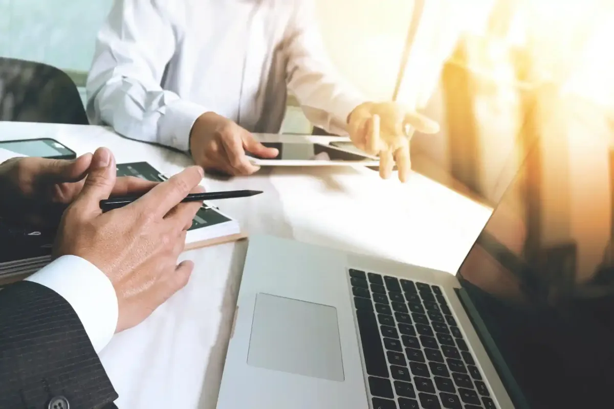 Due uomini d'affari seduti a un tavolo con computer portatile, tablet e smartphone; uno fa un gesto verso l'altro con i documenti.