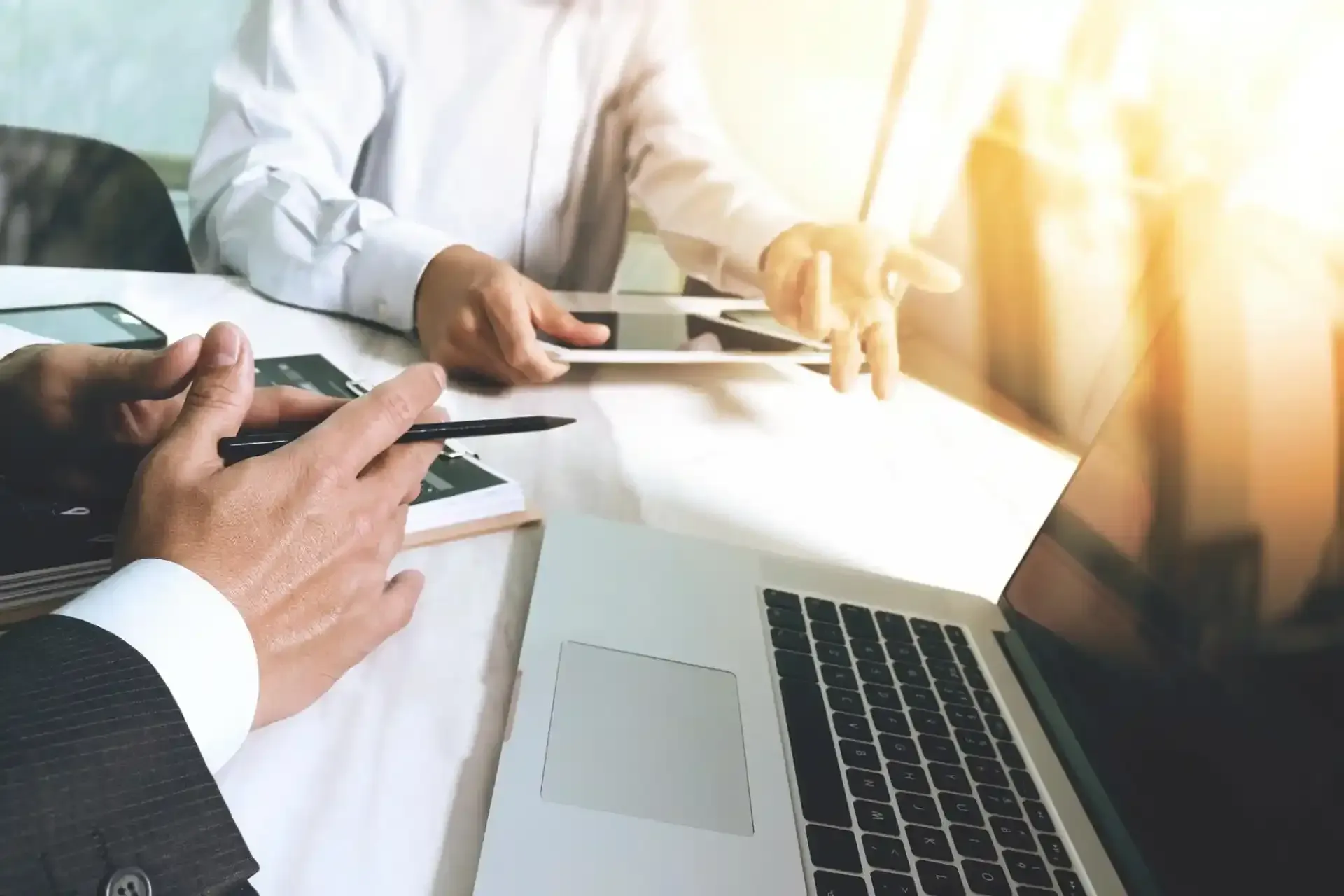 Due uomini d'affari seduti a un tavolo con computer portatile, tablet e smartphone; uno fa un gesto verso l'altro con i documenti.