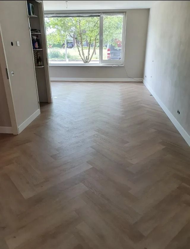 Lege kamer met lichte houten vloer in visgraatpatroon en groot raam met uitzicht op de bomen buiten.