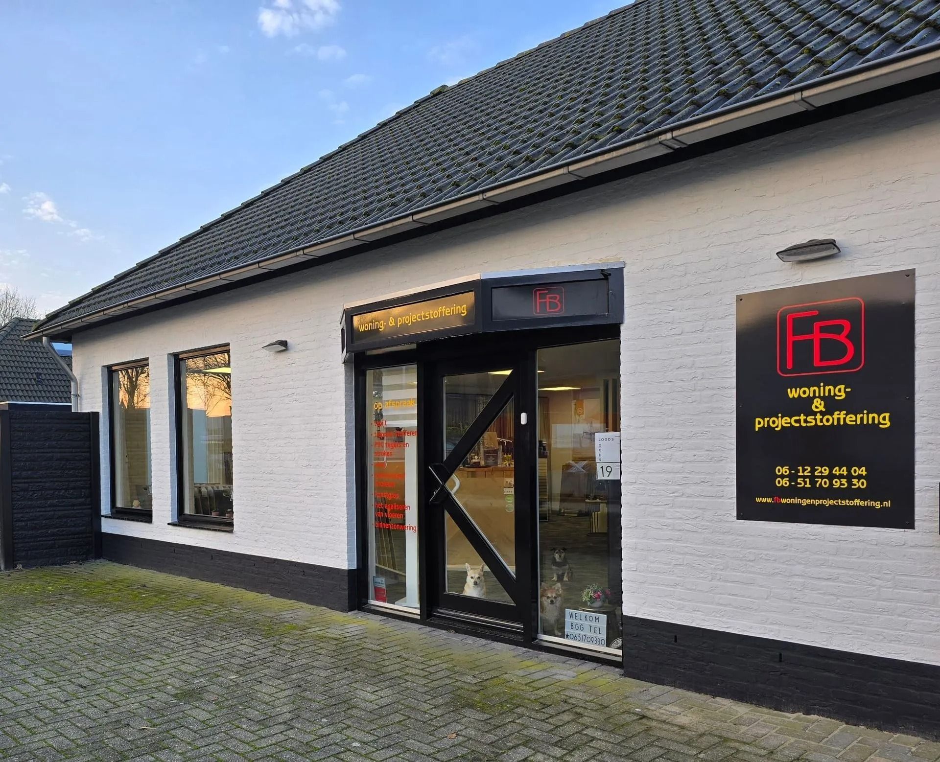 De buitenkant van een kleine winkelpui met witte muren, zwarte kozijnen en een rood-geel uithangbord bij de deur.