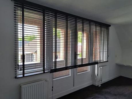 Lege kamer met grote zwarte jaloezieën voor de ramen en witte radiatoren eronder.