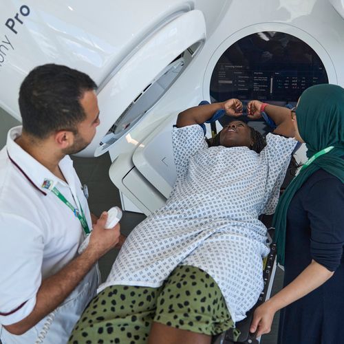 A medical professional and an assistant tend to a patient lying on a table in front of a medical imaging machine.