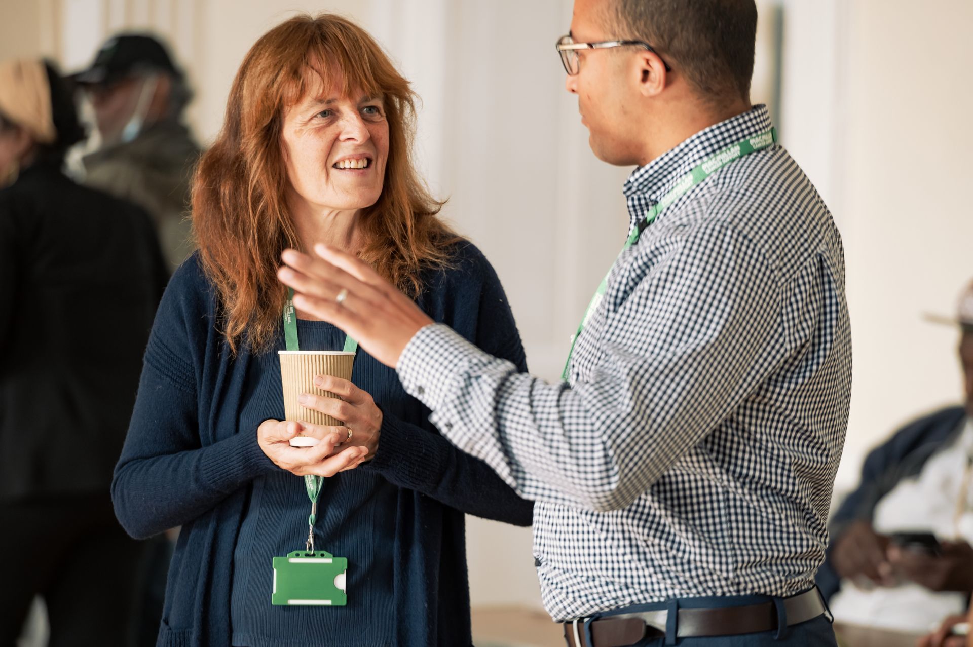 Two people in professional attire engage in a conversation, with one person gesturing while holding a coffee cup.