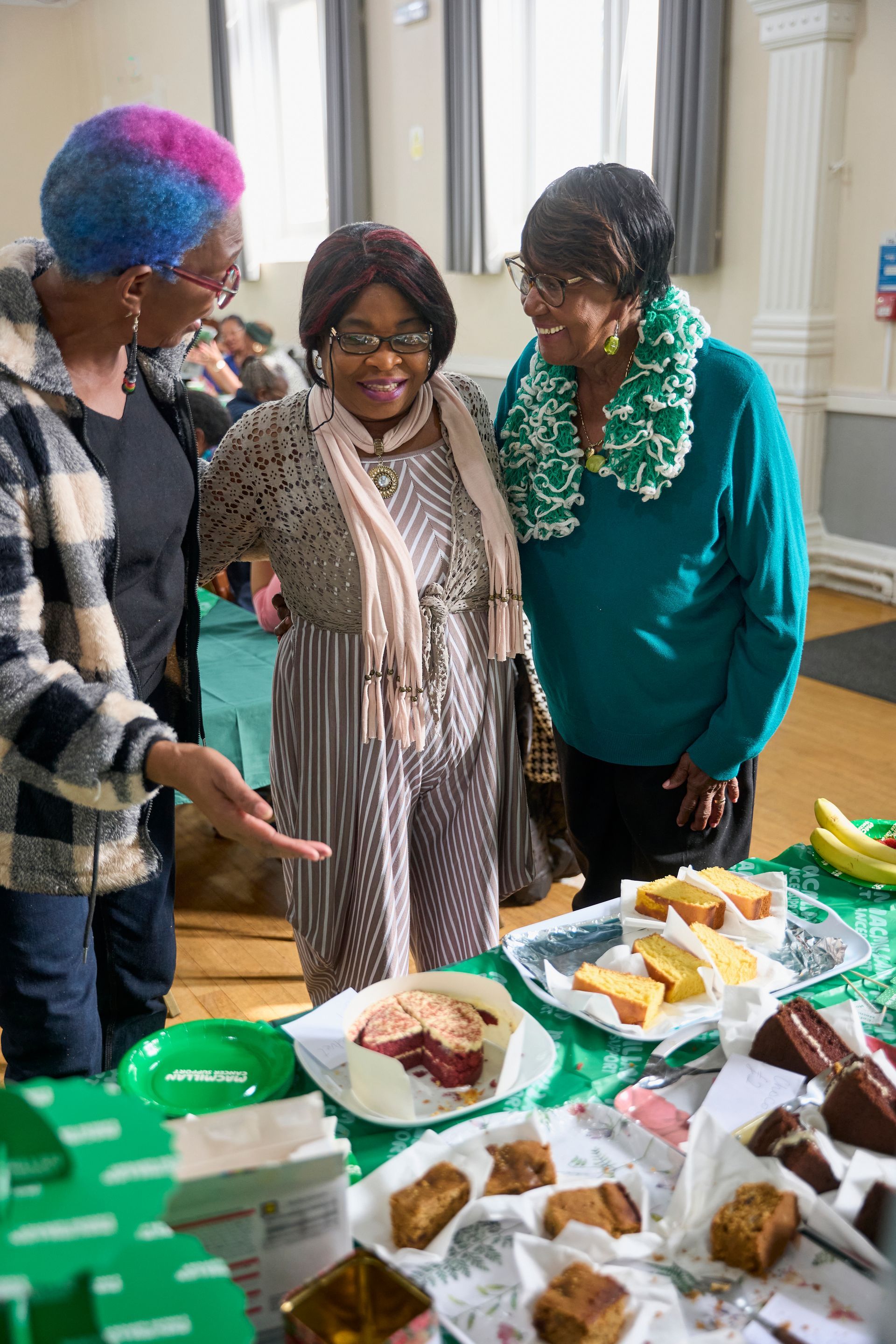 Three people stand around a table filled with various sliced cakes at a social gathering.