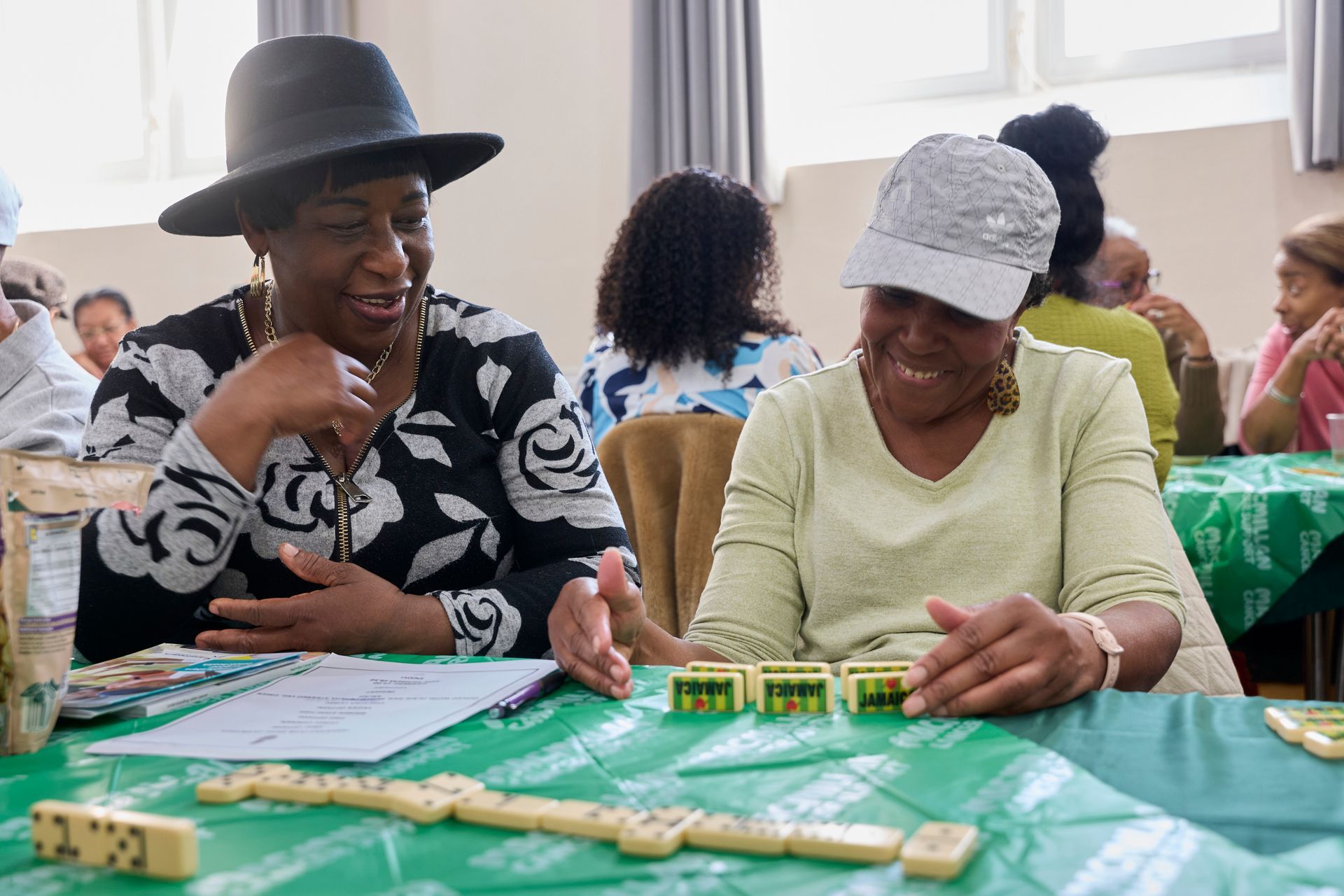 Two people wearing hats sit at a table covered in a green cloth, smiling while playing a game of dominoes.