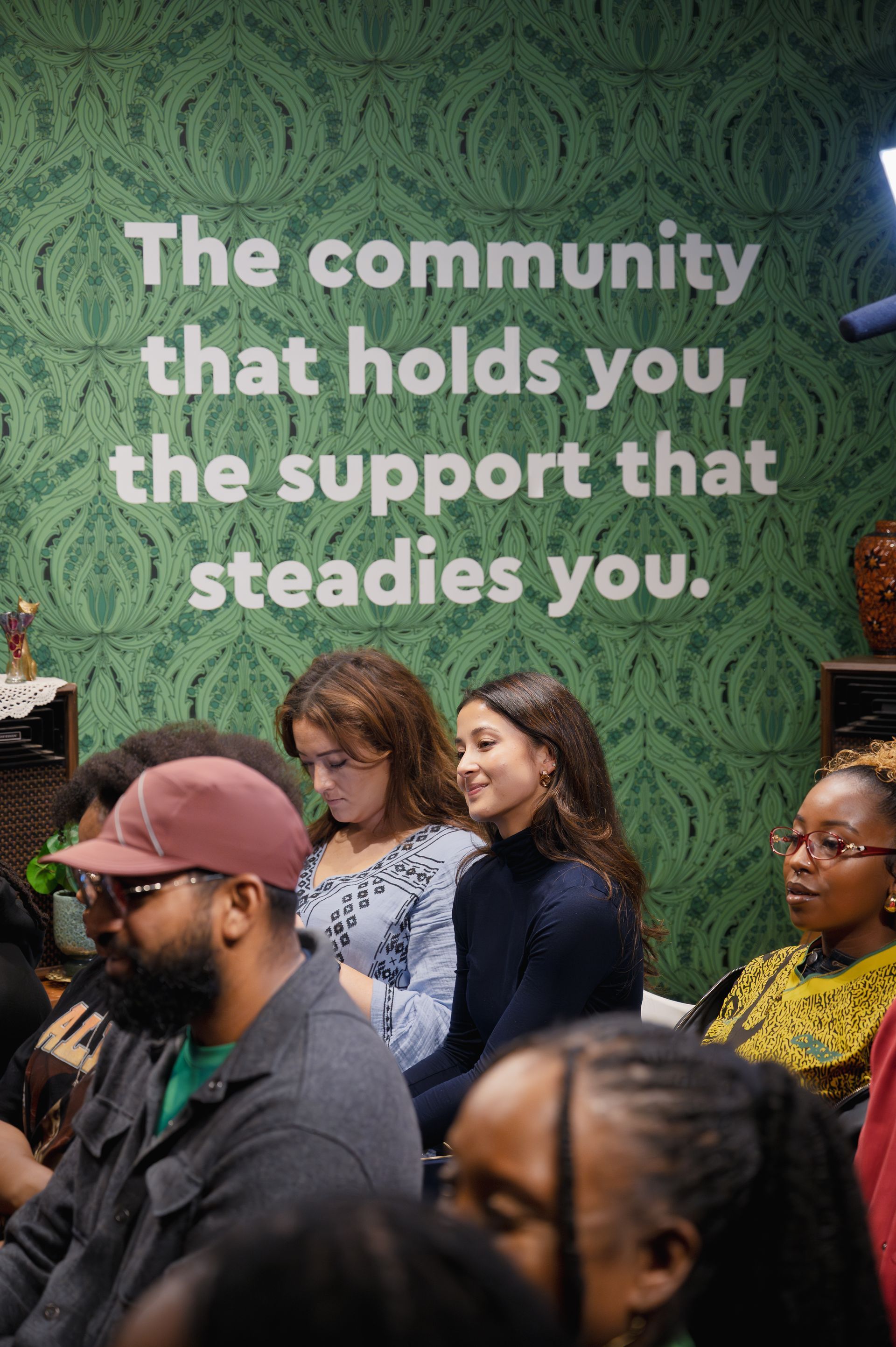 A group of people seated in a room with a green, patterned wall bearing the text, 