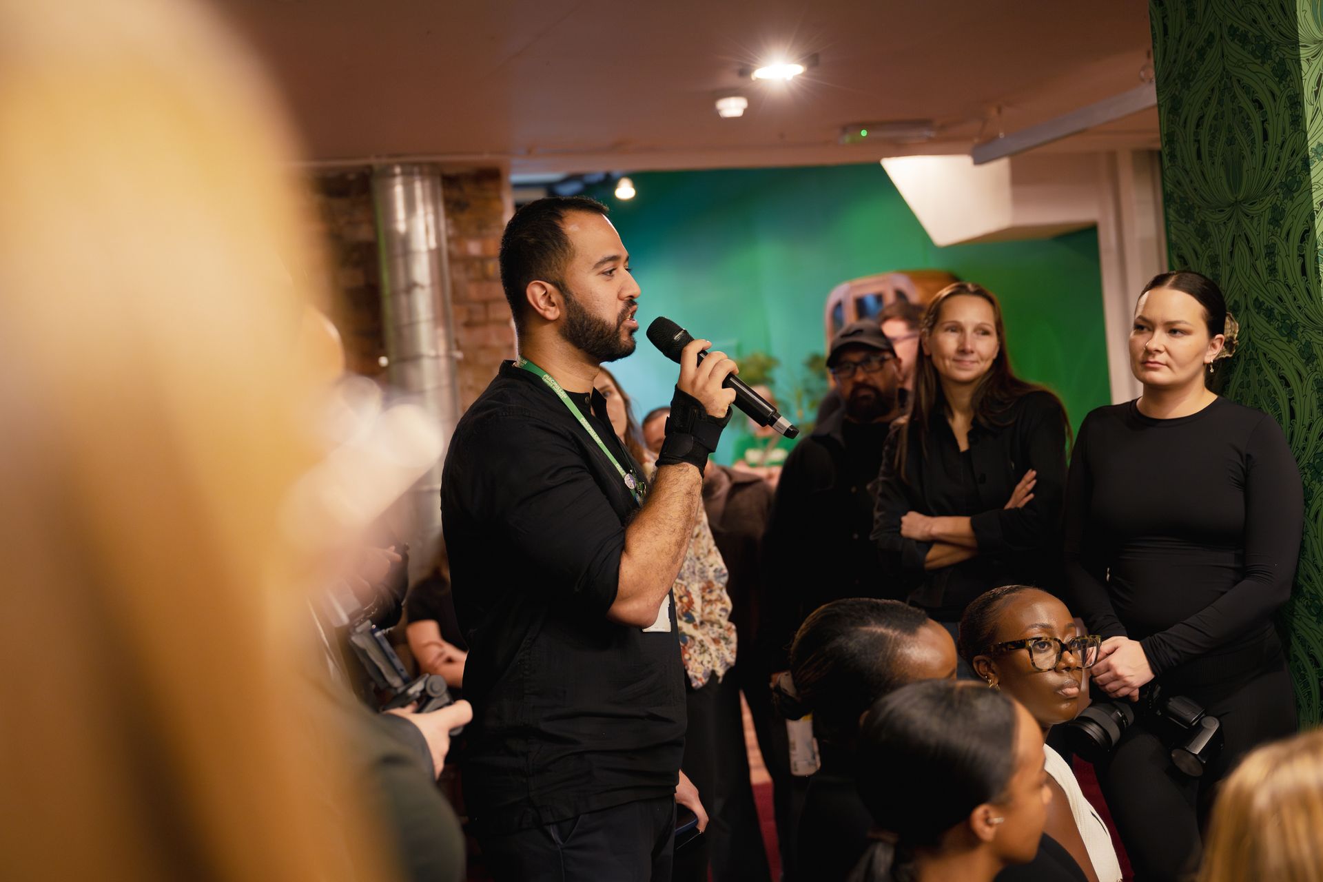 A person speaks into a microphone while standing before an attentive audience in a room with a green wall.