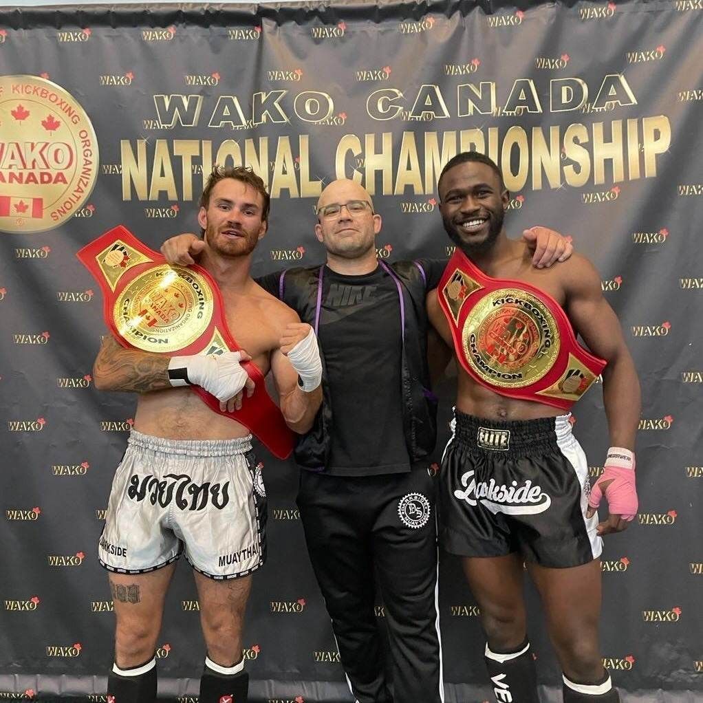 Three people posing with championship belts. Two athletes on either side of a coach. WAKO Canada banner in background.