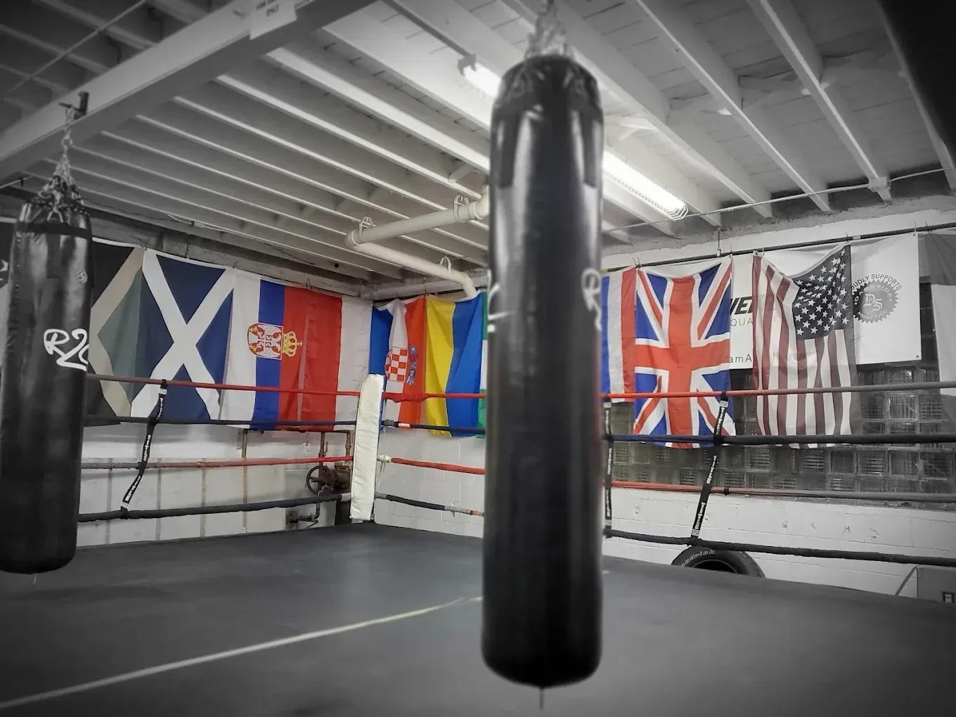 Boxing gym with heavy bag in front of boxing ring, flags of various nations hang on the wall.