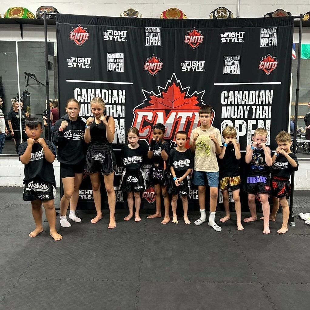 Group of Muay Thai fighters, young and adult, posing in a gym; black, red, white, and green colors.