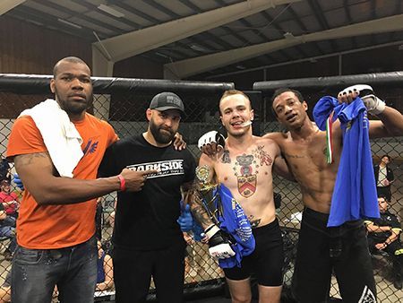 Four men pose in a boxing ring: a champion with a belt and two men, one with a towel, the other holding a blue shirt.