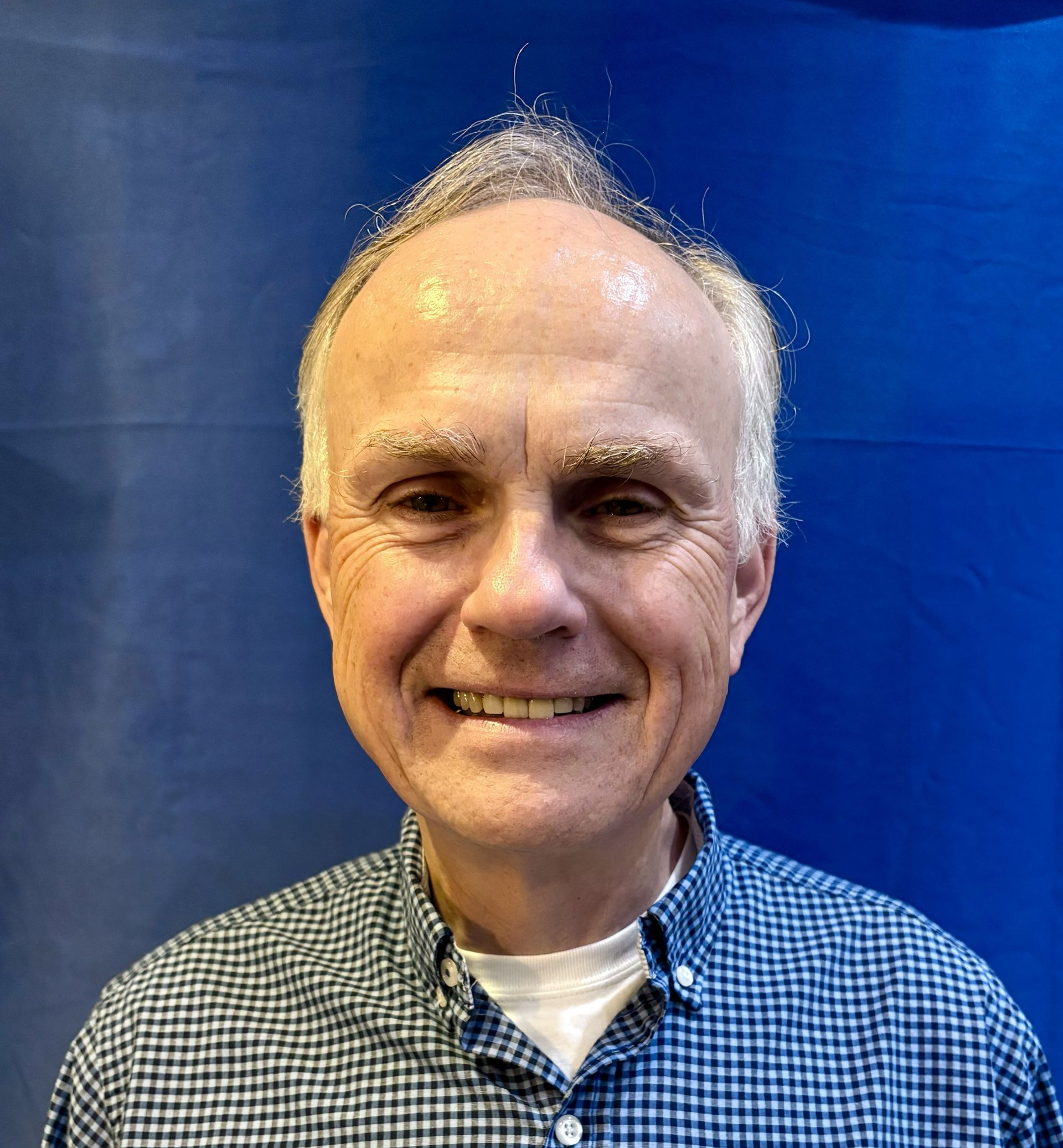 Man with gray hair and a blue checkered shirt smiles against a blue backdrop.