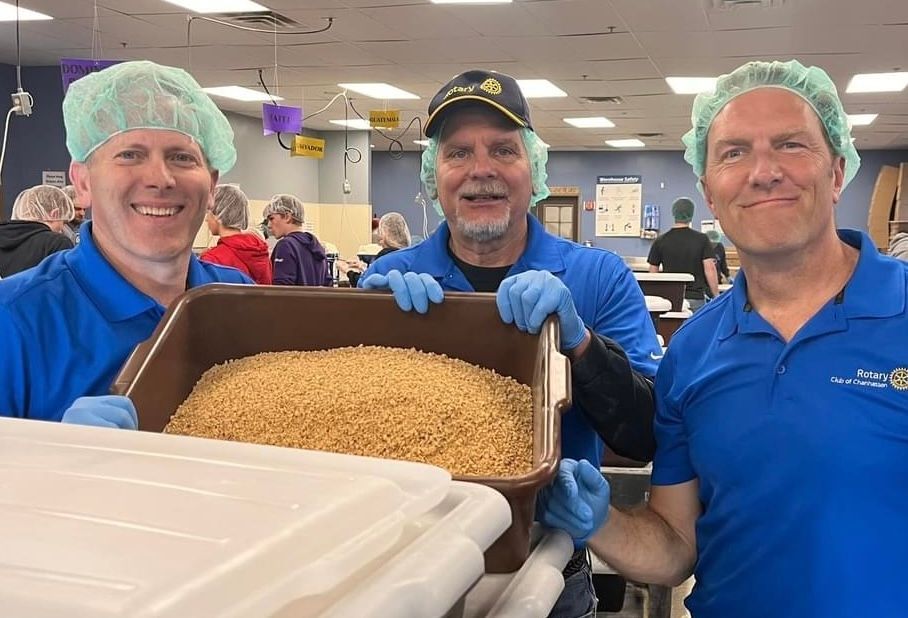 Three people in blue shirts, hairnets, and gloves holding a bin of grains. People in background.