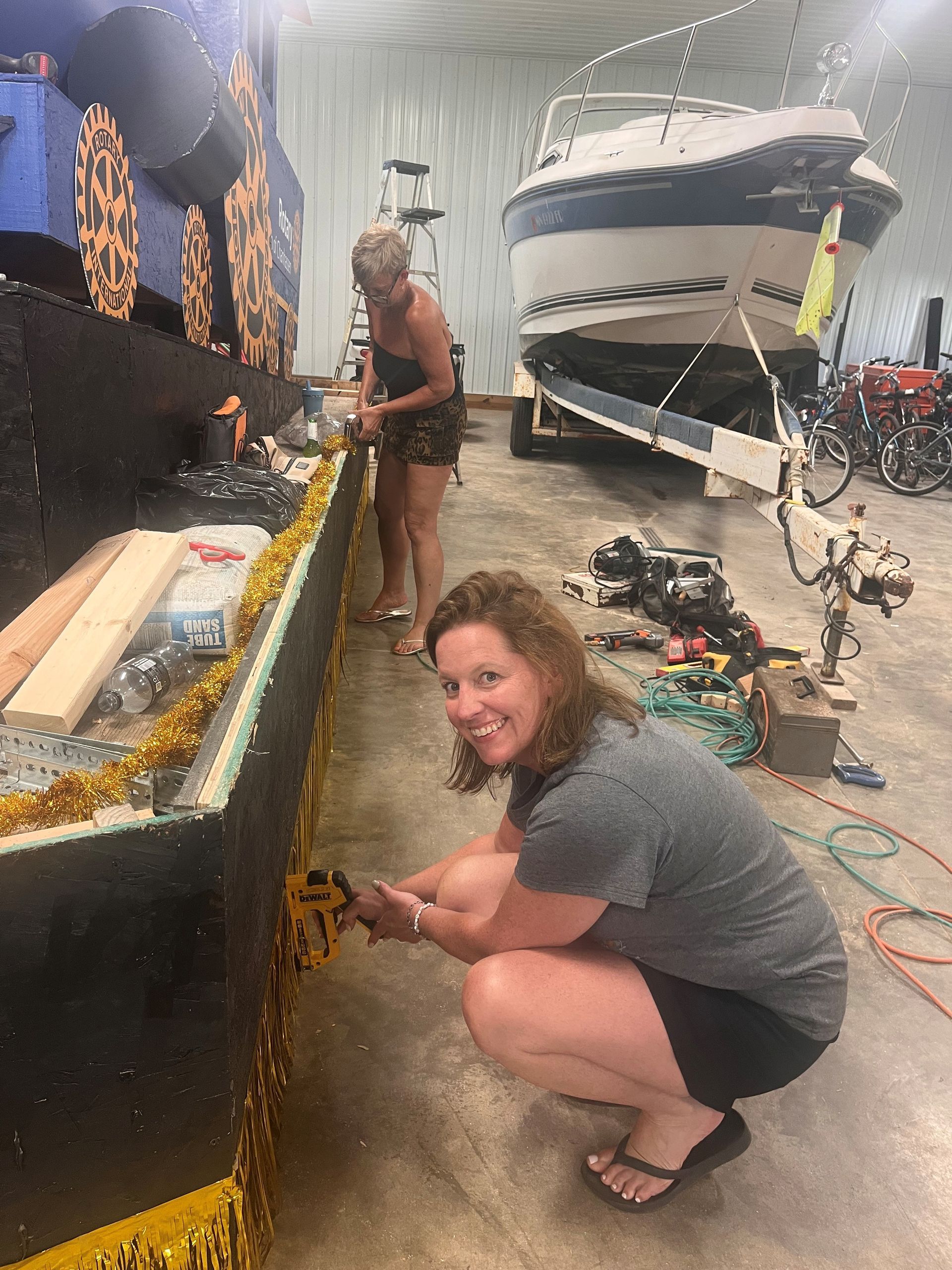 Two women decorating a float with gold trim inside a garage. One squats and smiles. A boat is in the background.