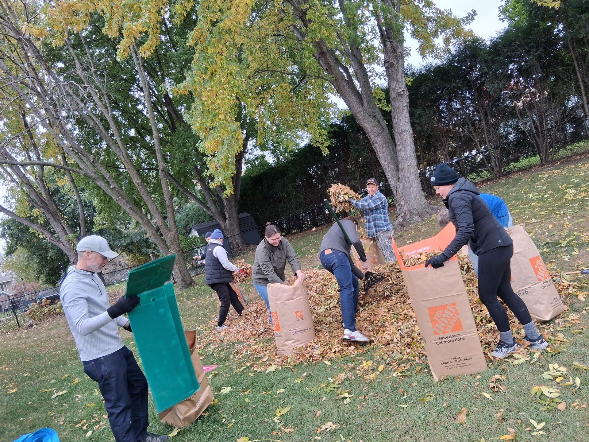 People raking and bagging leaves in a yard. Autumn setting, brown leaves, paper bags, some wearing jackets.