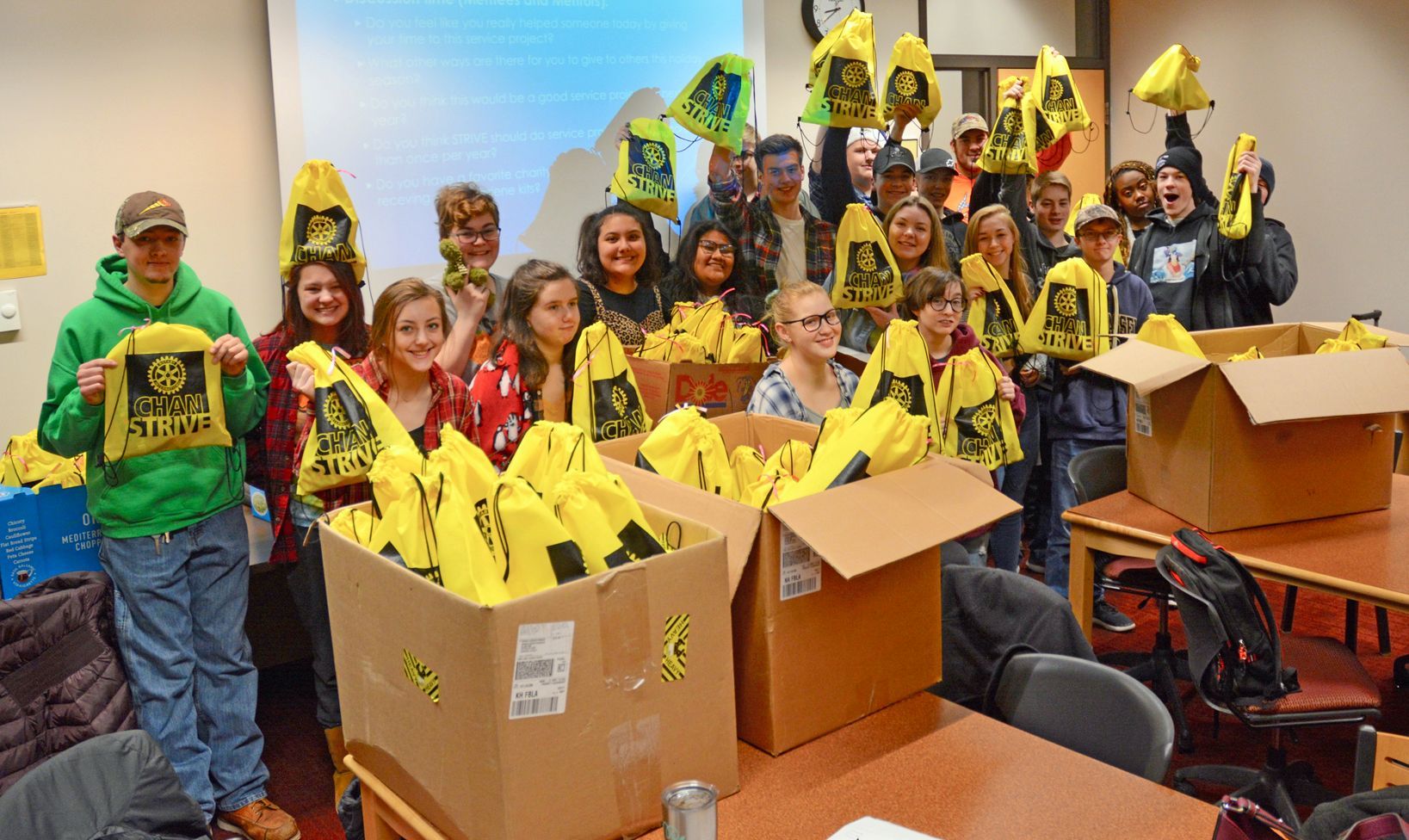 Group of people holding up yellow bags with a logo; indoors, smiling, near boxes of bags.
