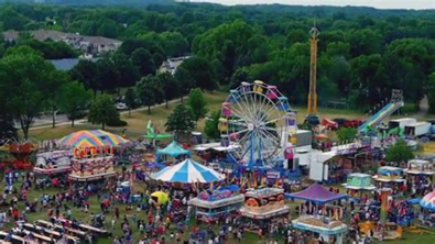 Aerial view of a busy outdoor fair with rides, food stands, and many people gathered on the grass.