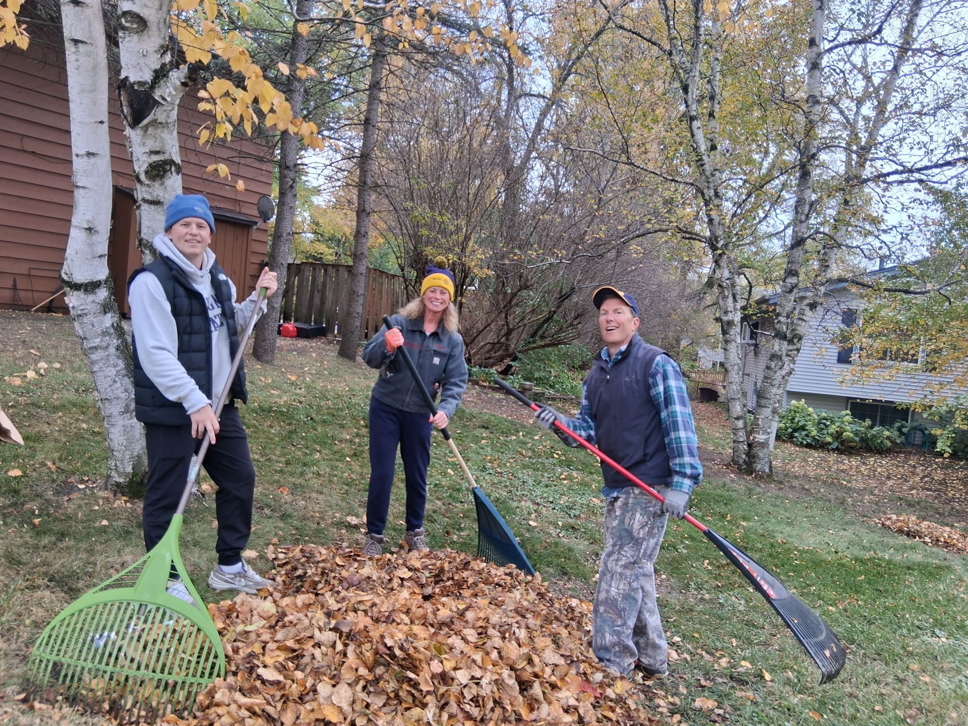Three people raking leaves outdoors: two women, one man. Fall foliage, overcast sky.
