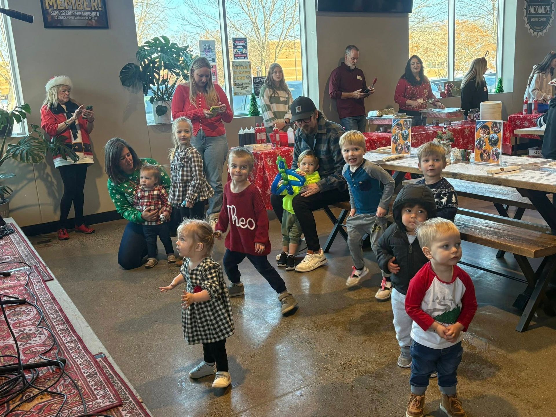 Children and adults gathered in a brightly lit room for an event. Several children are in front, some with Christmas attire.