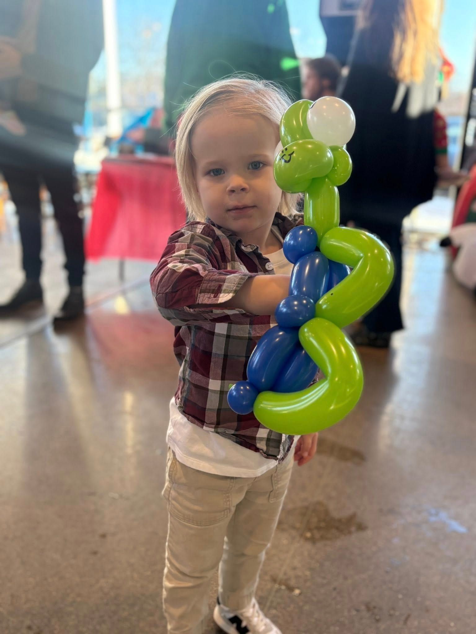 Young child holding a green and blue balloon animal; indoor setting with other people.