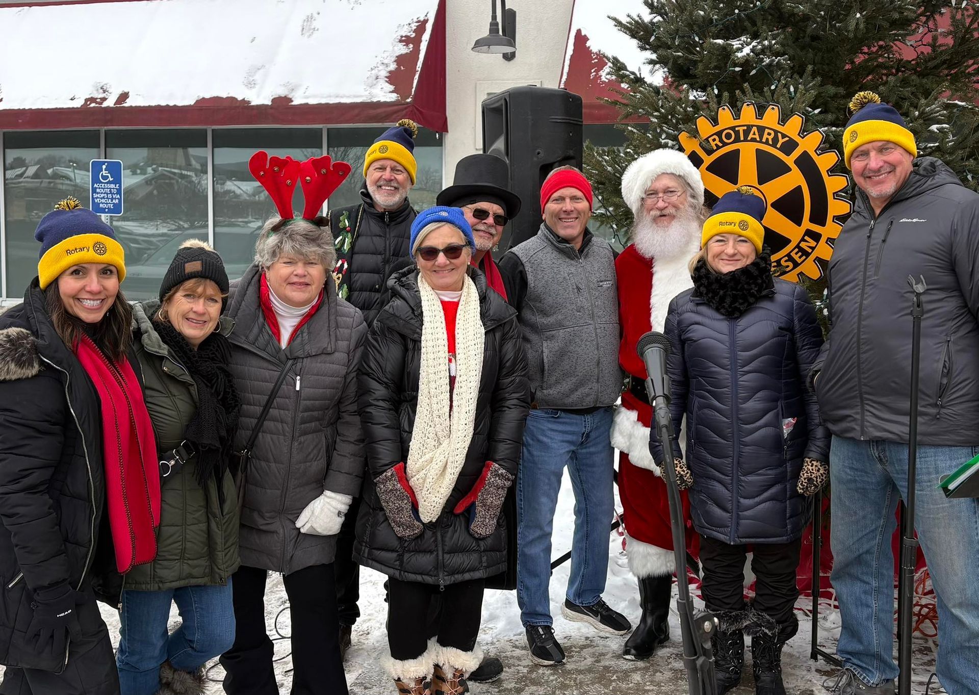 Group of people, including Santa, wearing yellow and blue hats, posing near a Rotary Club sign in a snowy outdoor setting.