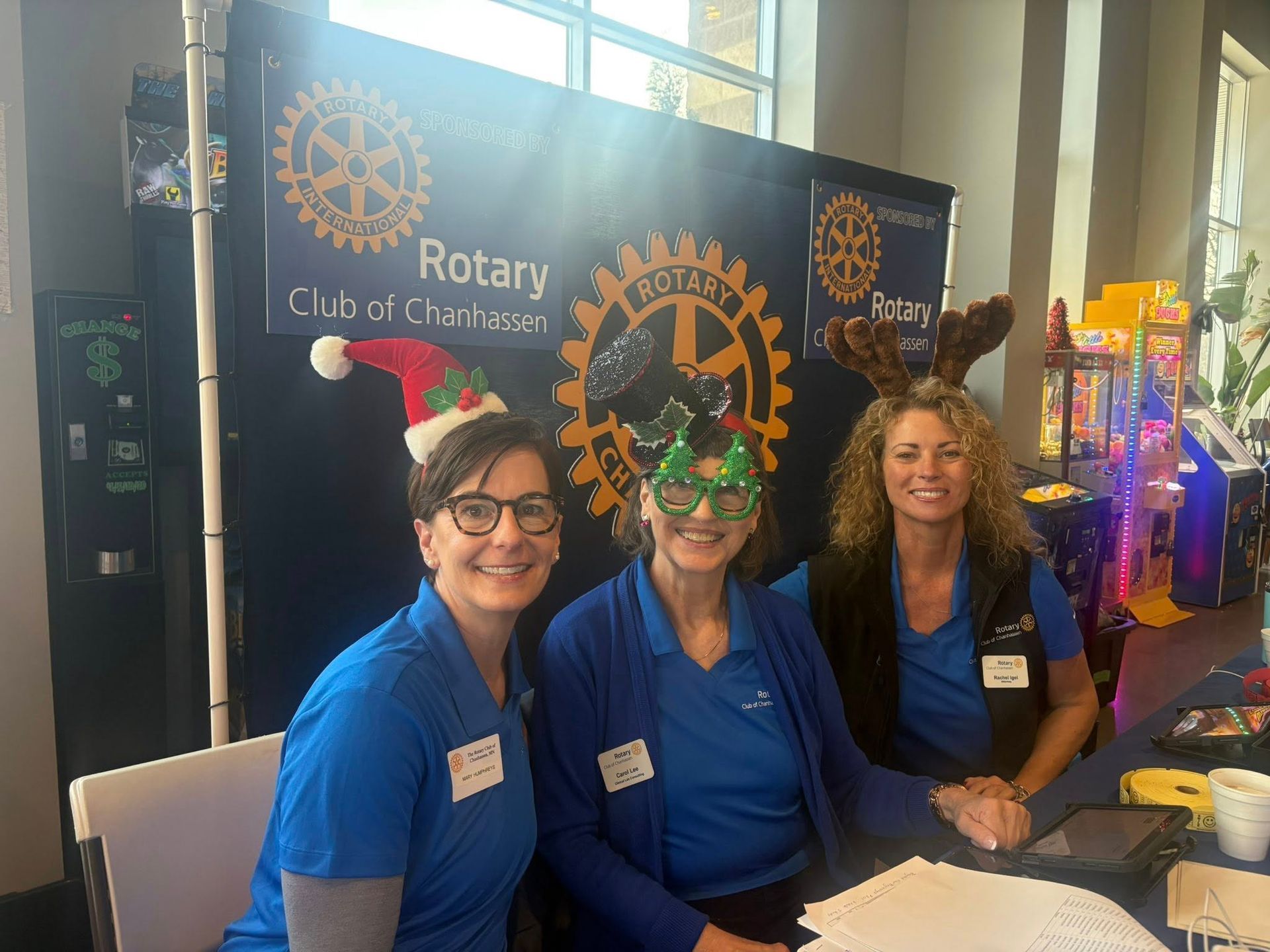 Three people in blue shirts stand behind a table at a Rotary Club event. They wear holiday-themed headgear, smiling.