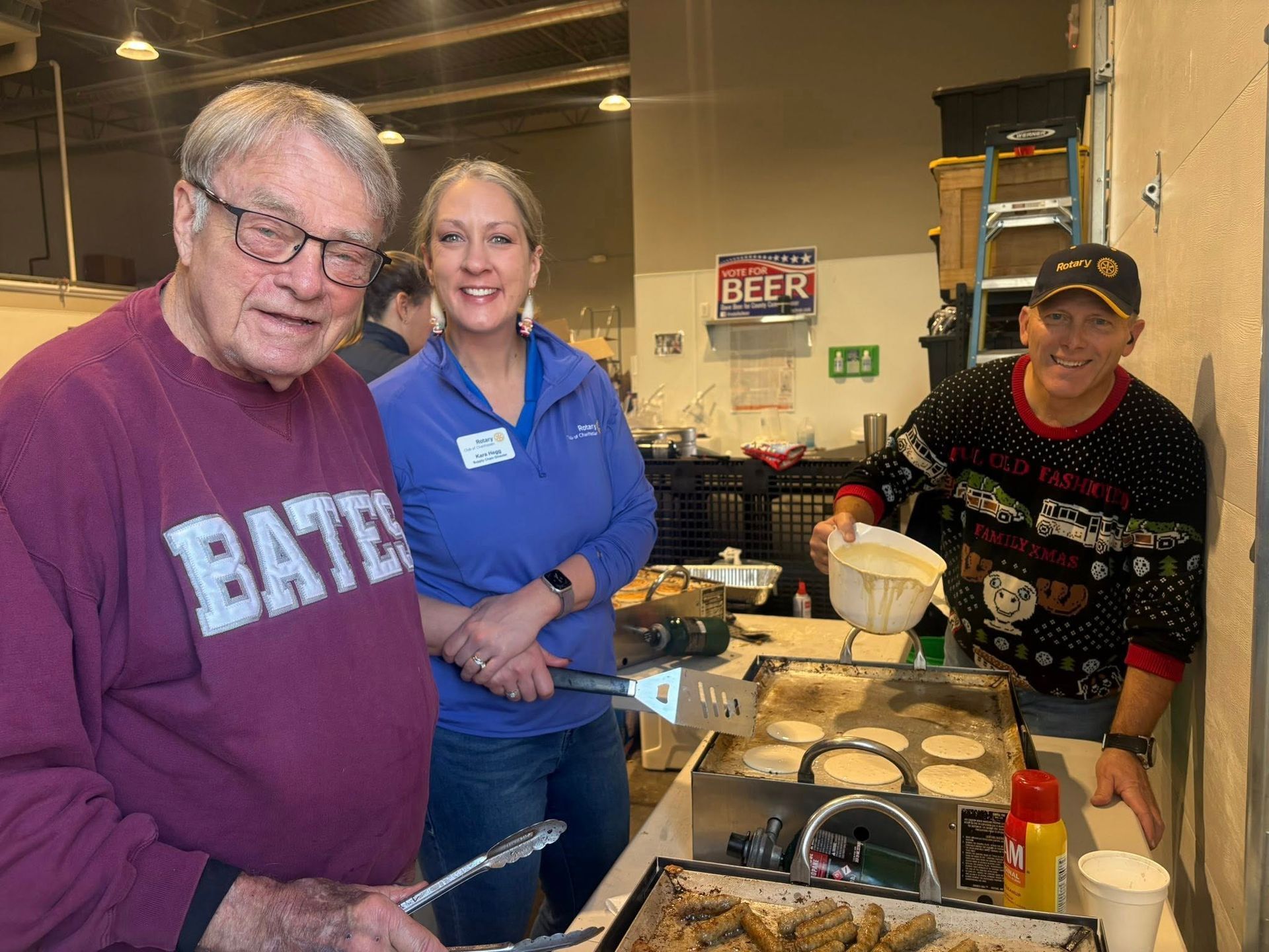 People at an event, one wearing a sweater pouring batter onto a griddle, others smiling, food on display.