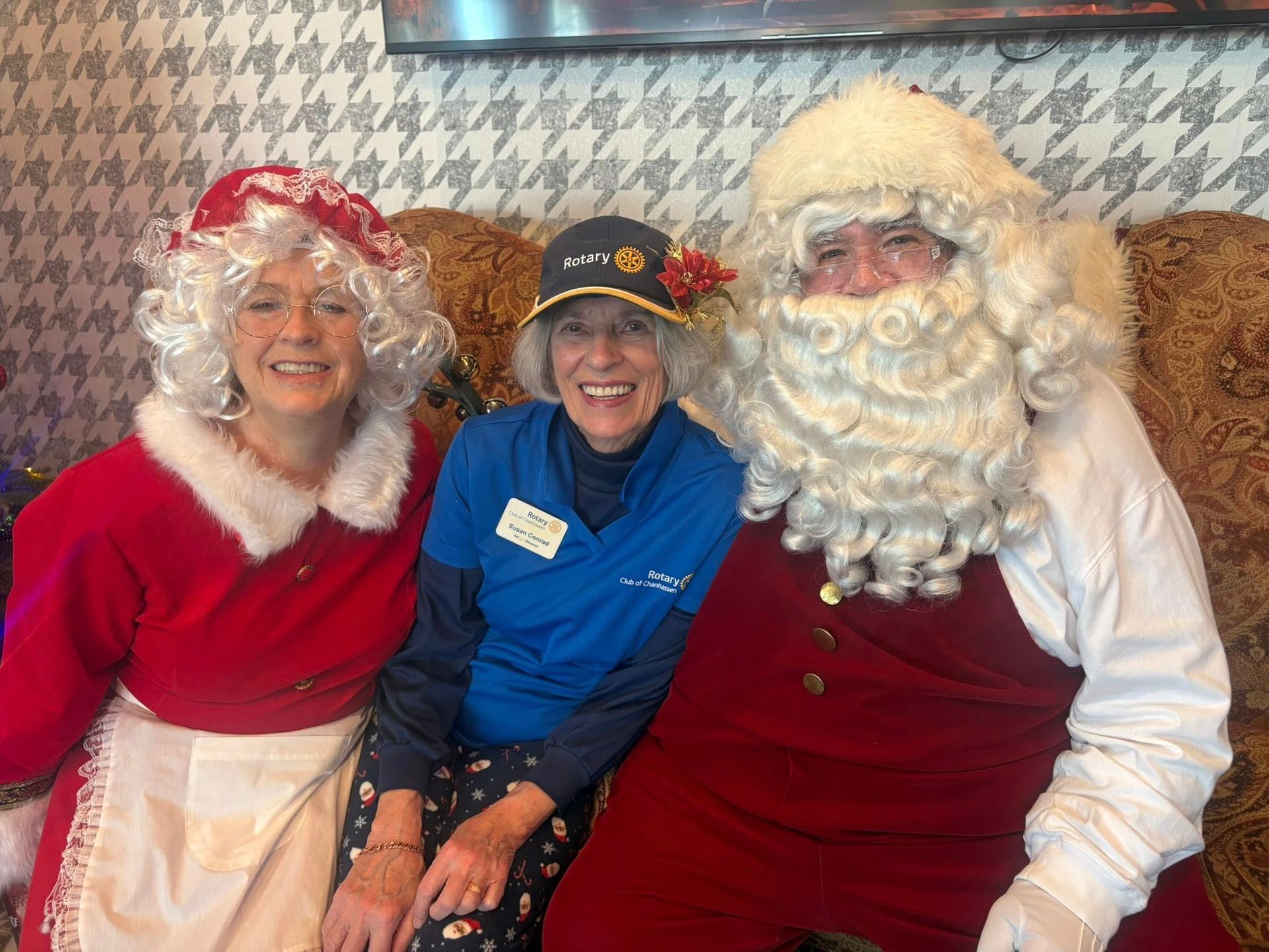 Mrs. Claus, a volunteer, and Santa Claus sit on a couch. They're smiling in front of a patterned wall.