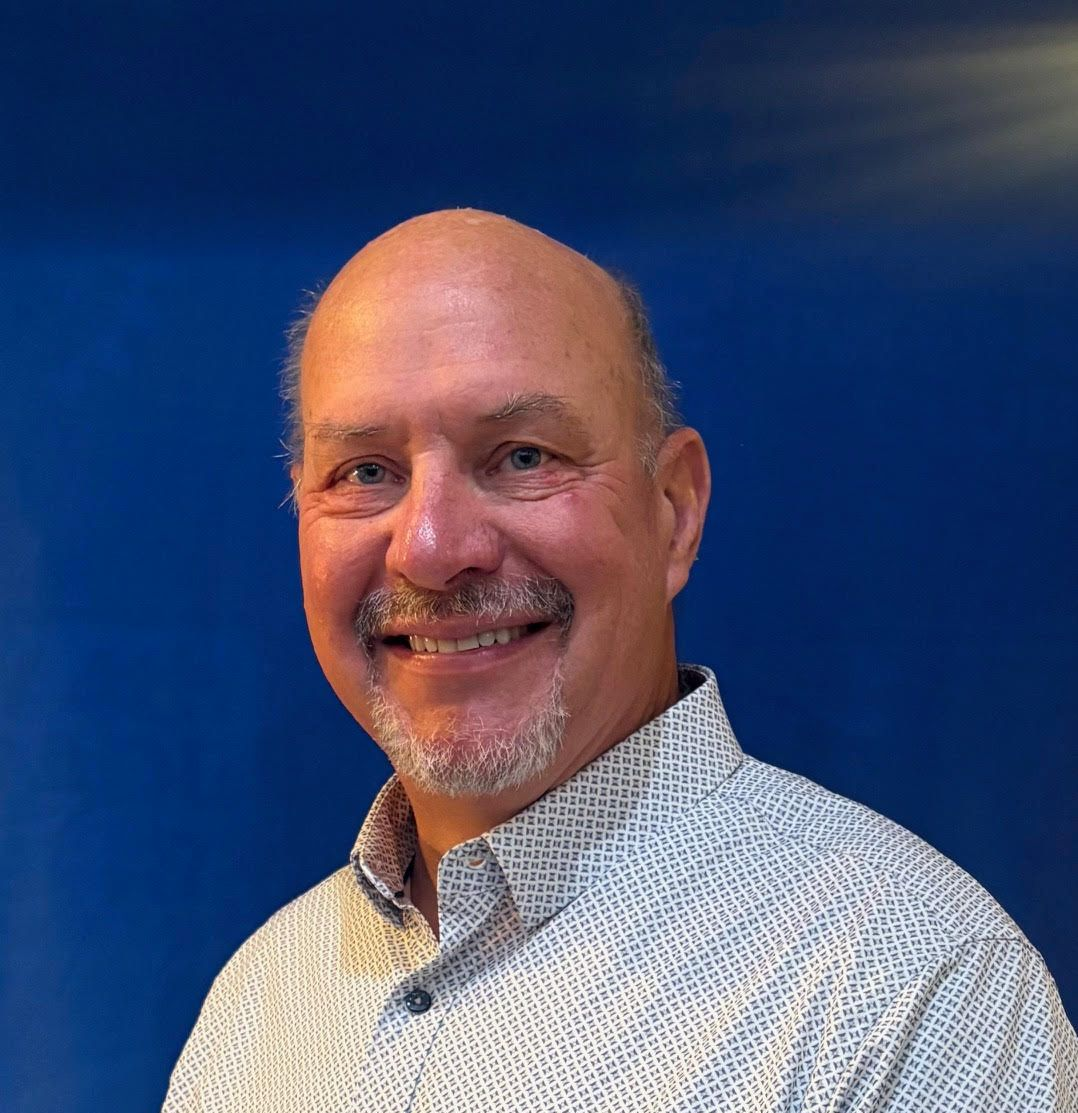 Bald man with a graying goatee smiles, wearing a patterned white and blue button-down shirt. Blue background.