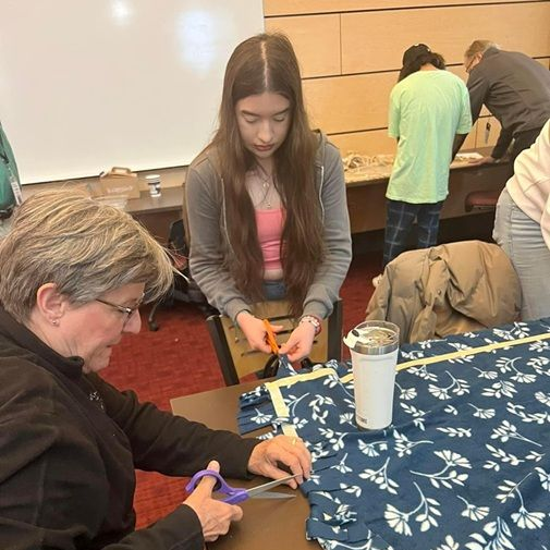 Two people cutting fabric at a table; others in the background.