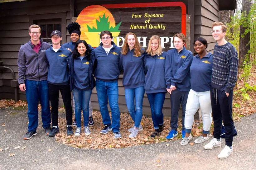 Group of people in matching jackets stand in front of a sign that reads "Wood Pile."